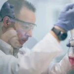 Scientist in lab coat and gloves transferring pink liquid using pipette.